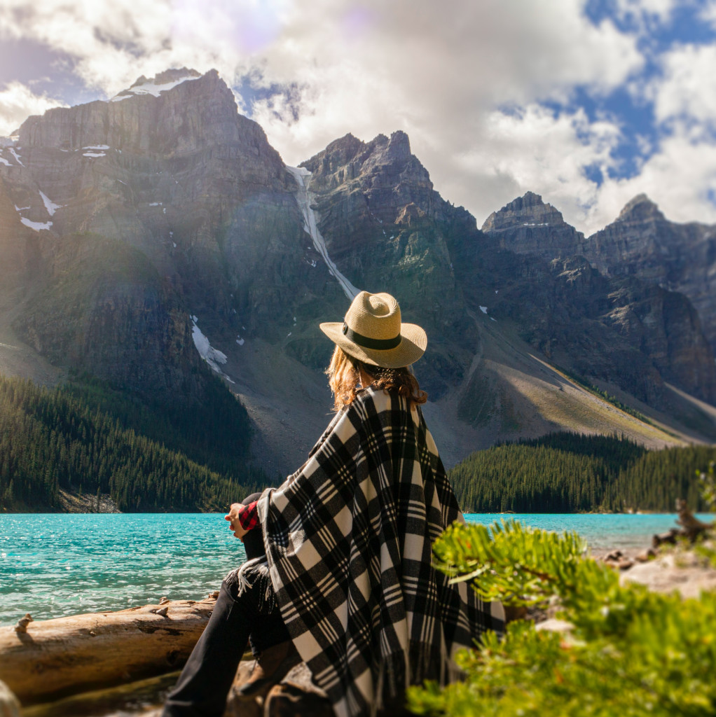 A woman sits beside a vast lake gazing towards a vista of rocky mountains